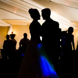 Silhouetted bride and groom dancing at a wedding reception, colorful lights illuminating the bottom of the dress, guests in the background.