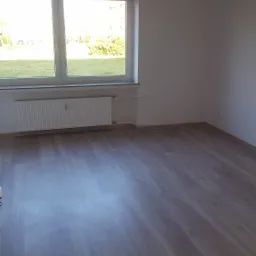 Puste, jasne pomieszczenie z panelami podłogowymi, białymi ścianami, grzejnikiem pod oknem i wiszącą żarówką bez klosza.