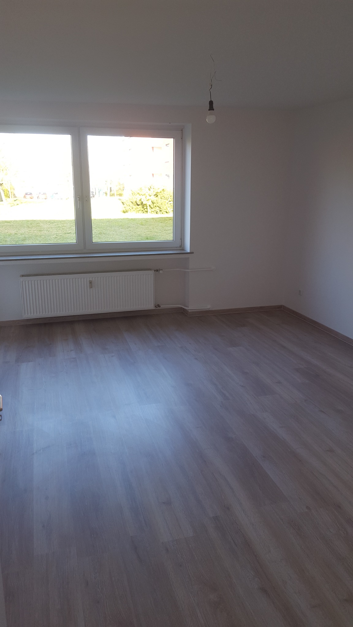 Puste, jasne pomieszczenie z panelami podłogowymi, białymi ścianami, grzejnikiem pod oknem i wiszącą żarówką bez klosza.