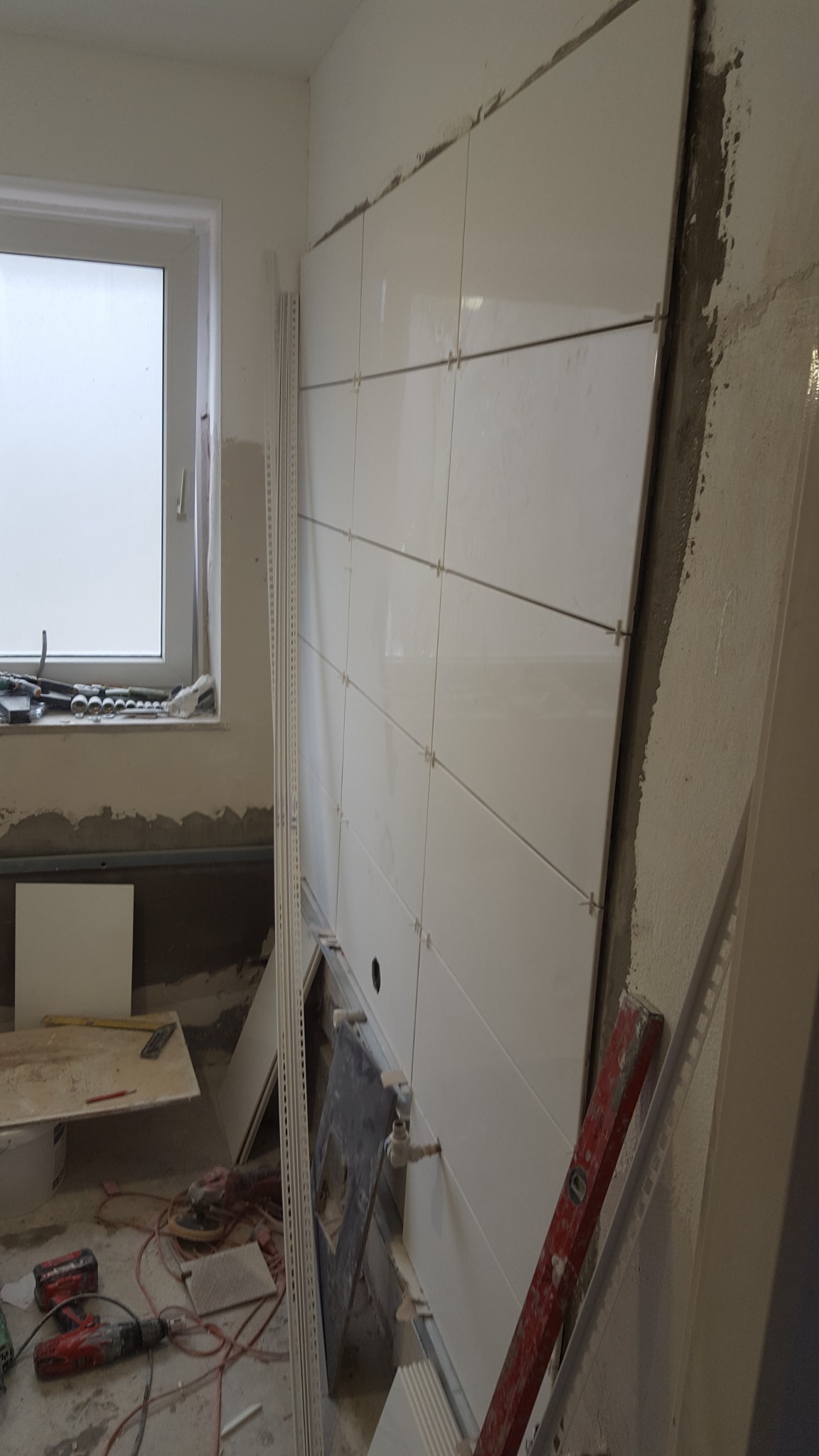 Ściana łazienki w trakcie układania białych płytek, widoczne krzyżyki dystansowe, poziomica i narzędzia na podłodze, przygotowane miejsce pod umywalkę.