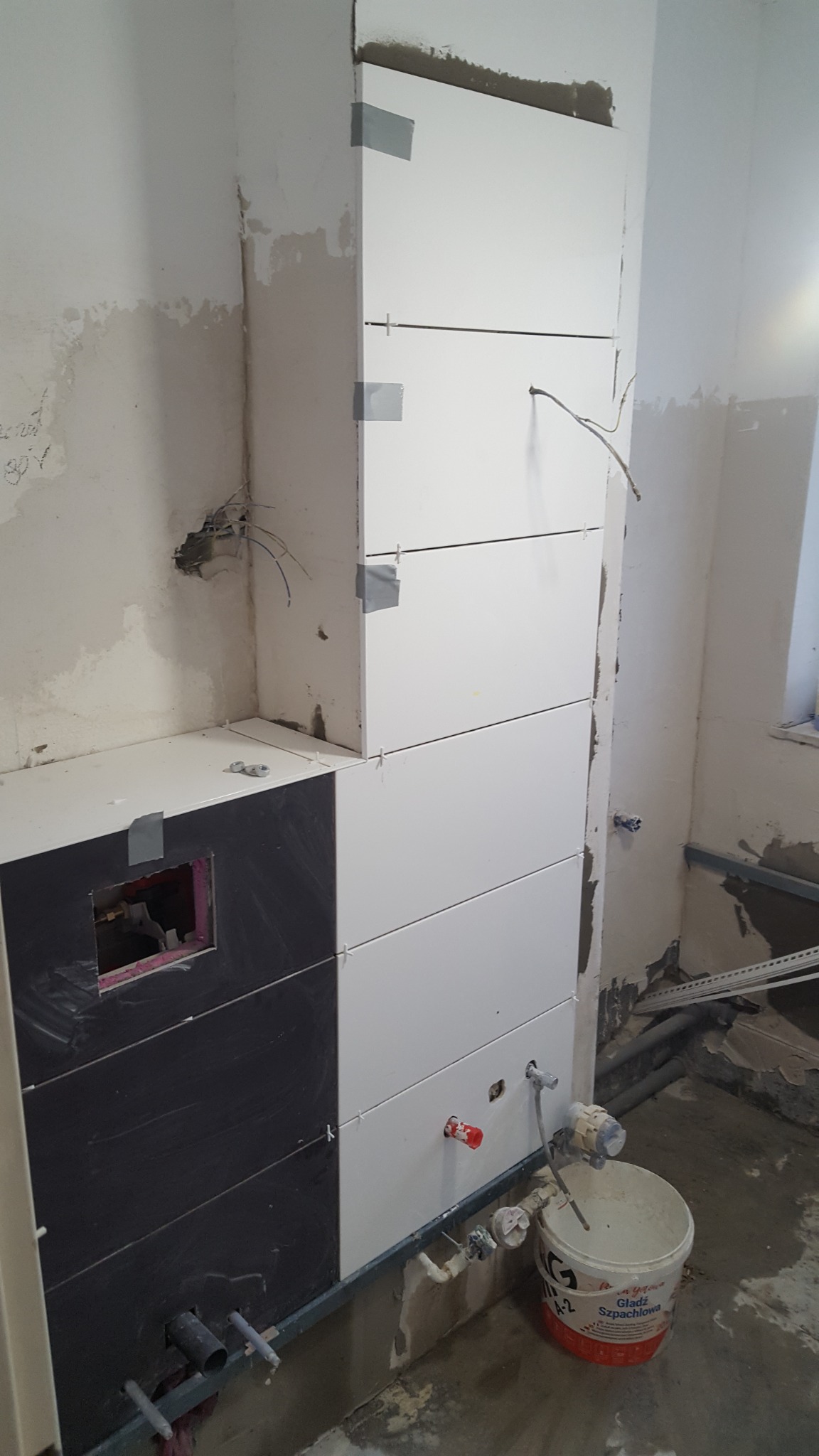 Wnętrze w trakcie remontu: częściowo wyłożona biało-czarnymi płytkami ściana z widocznymi instalacjami hydraulicznymi i elektrycznymi, zabezpieczona taśmą malarską, z wiadrem po gładzi szpachlowej...