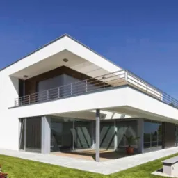 Nowoczesny dom z białymi ścianami, dużymi przeszkleniami i balkonem z metalową barierką, otoczony zielonym trawnikiem i roślinnością.