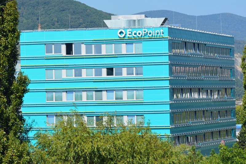 Turkusowy biurowiec EcoPoint z licznymi białymi oknami, widok z dołu na tle zalesionych wzgórz.