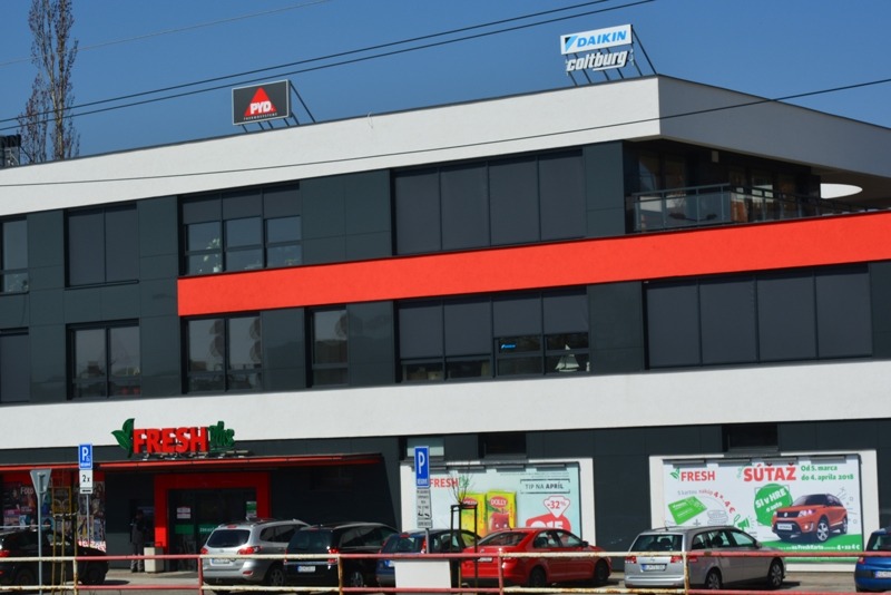 Dwupiętrowy budynek biurowo-usługowy z ciemnymi oknami, czerwonym pasem na elewacji i szyldami reklamowymi na dachu, widziany z poziomu ulicy z zaparkowanymi samochodami.