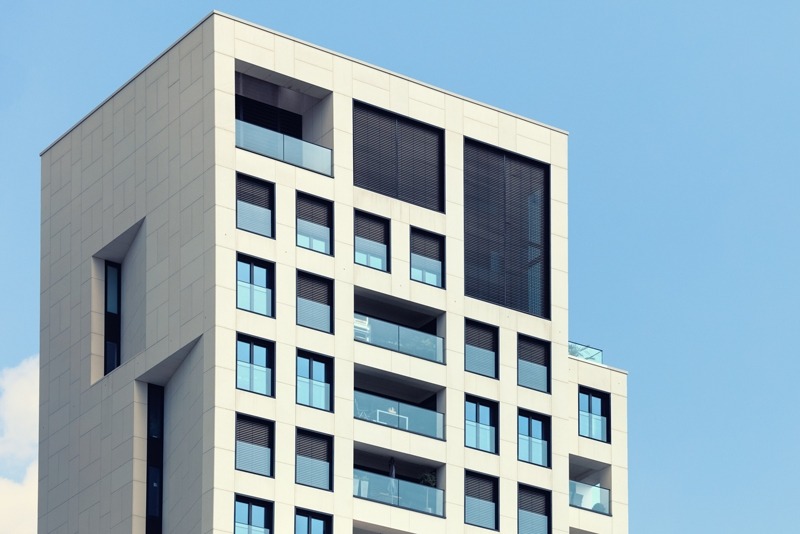 Fragment nowoczesnego budynku z jasną elewacją, regularnie rozmieszczonymi oknami z ciemnymi ramami i szklanymi balustradami balkonów, na tle błękitnego nieba.