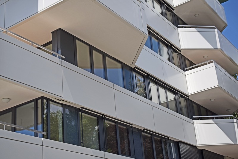 Nowoczesny budynek mieszkalny z balkonami i dużymi oknami w czarnych ramach, widok z dołu w słoneczny dzień.