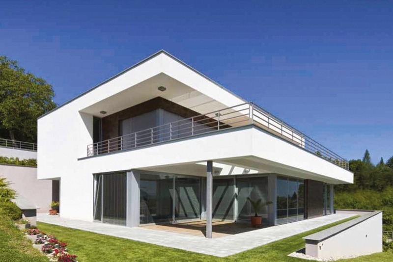 Nowoczesny dom z białymi ścianami, dużymi przeszkleniami i balkonem z metalową barierką, otoczony zielonym trawnikiem i roślinnością.