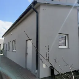 Szary budynek z nową elewacją, widoczne okna z białymi ramami, rynna spustowa i chodnik z kostki brukowej. W tle błękitne niebo z lekkimi chmurami.