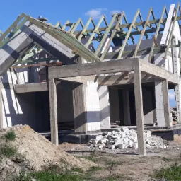 Surowy stan domu z białych bloczków, widoczne drewniane elementy konstrukcji dachu, betonowe filary i fundamenty, gruz budowlany przed budynkiem