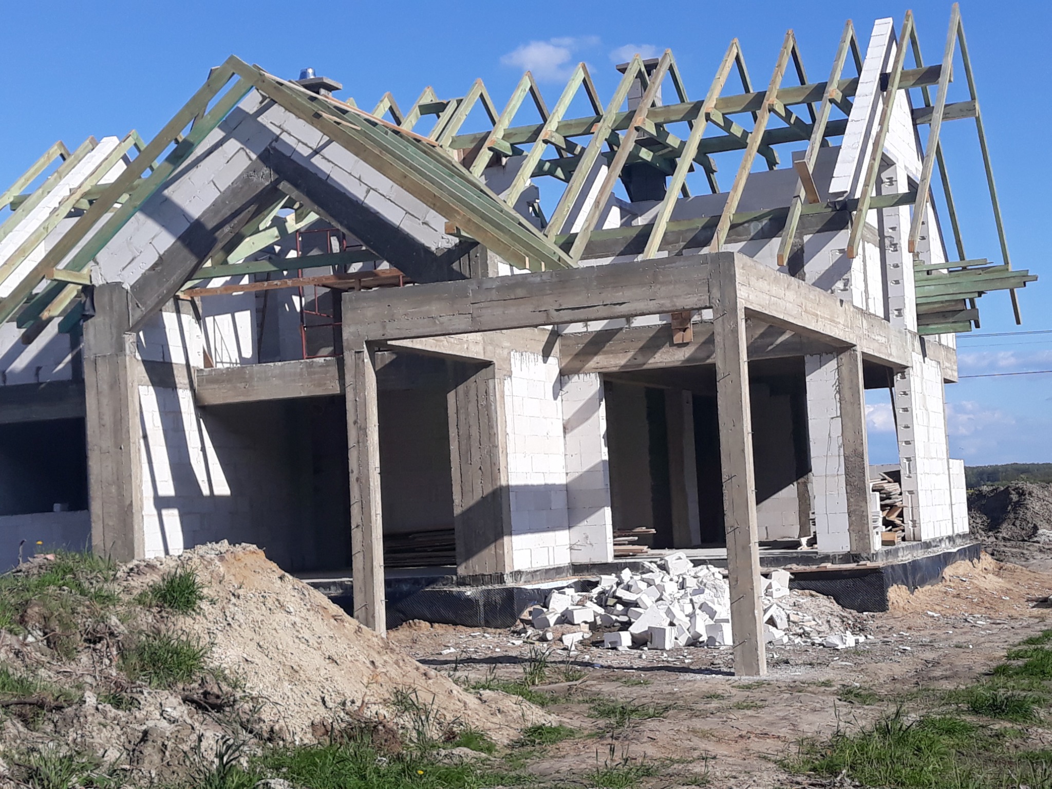 Surowy stan domu z białych bloczków, widoczne drewniane elementy konstrukcji dachu, betonowe filary i fundamenty, gruz budowlany przed budynkiem