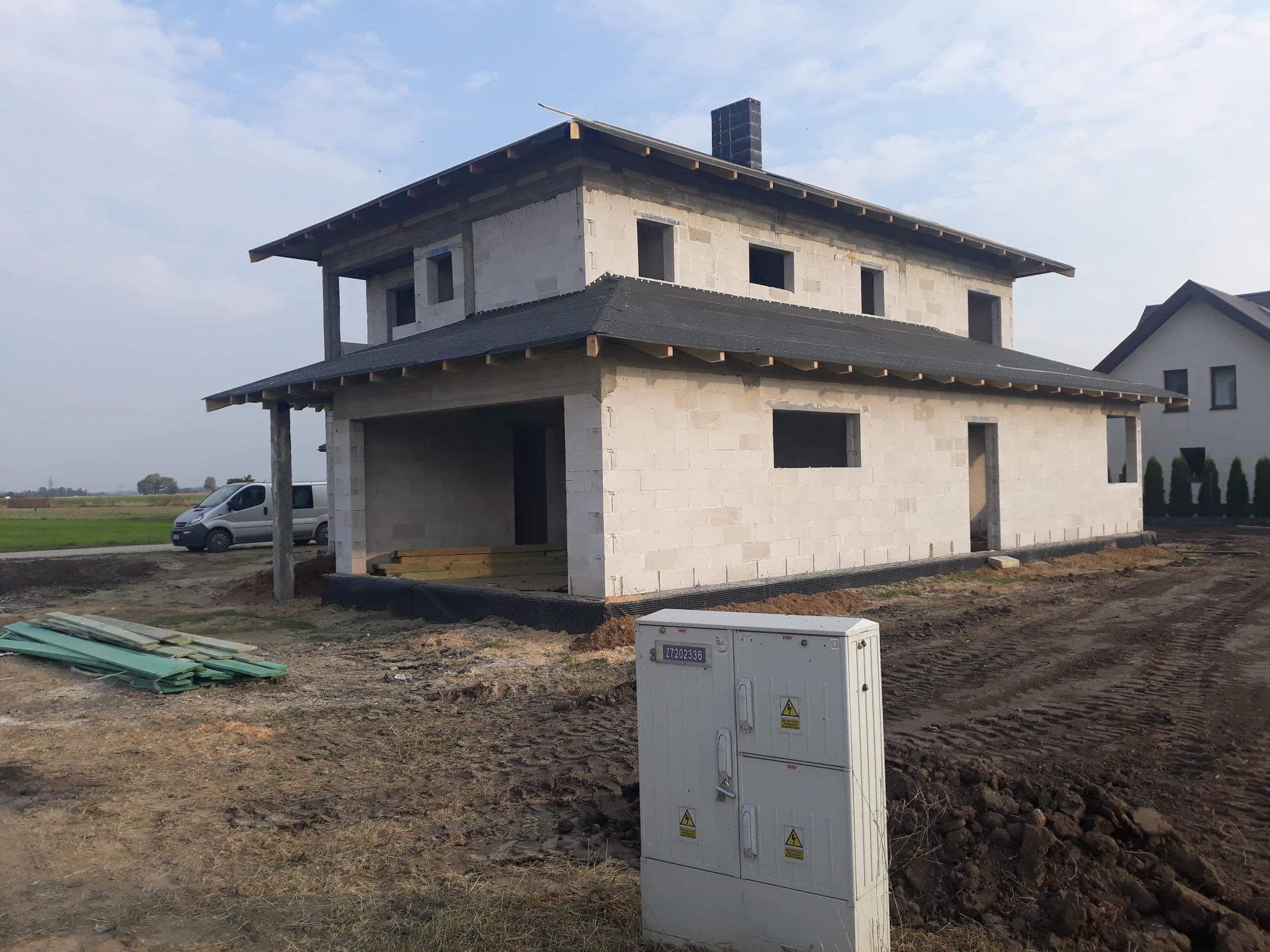 Niedokończony dwukondygnacyjny dom z białego bloczka, z widocznym zadaszeniem i kominem, otoczony rozkopanym terenem i skrzynką elektryczną, na tle błękitnego nieba.