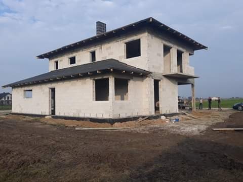 Surowy stan dwukondygnacyjnego domu z betonu komórkowego z widocznym dachem i kominem, w tle grupa osób.