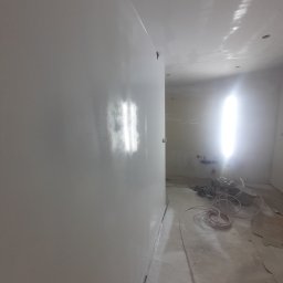 KJ Wykończenia Wnętrz - Świeżo pomalowane, białe, błyszczące ściany w pomieszczeniu. Widoczne narzędzia malarskie i niedoskonałości podłogi, sugerujące trwający remont. Oświetlenie halogenowe podkreśla gładkość powierzchni.