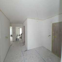 KJ Wykończenia Wnętrz - Białe ściany w mieszkaniu po szpachlowaniu, zabezpieczone podłogi folią malarską, widoczne kable wystające z sufitu, przygotowanie do malowania.