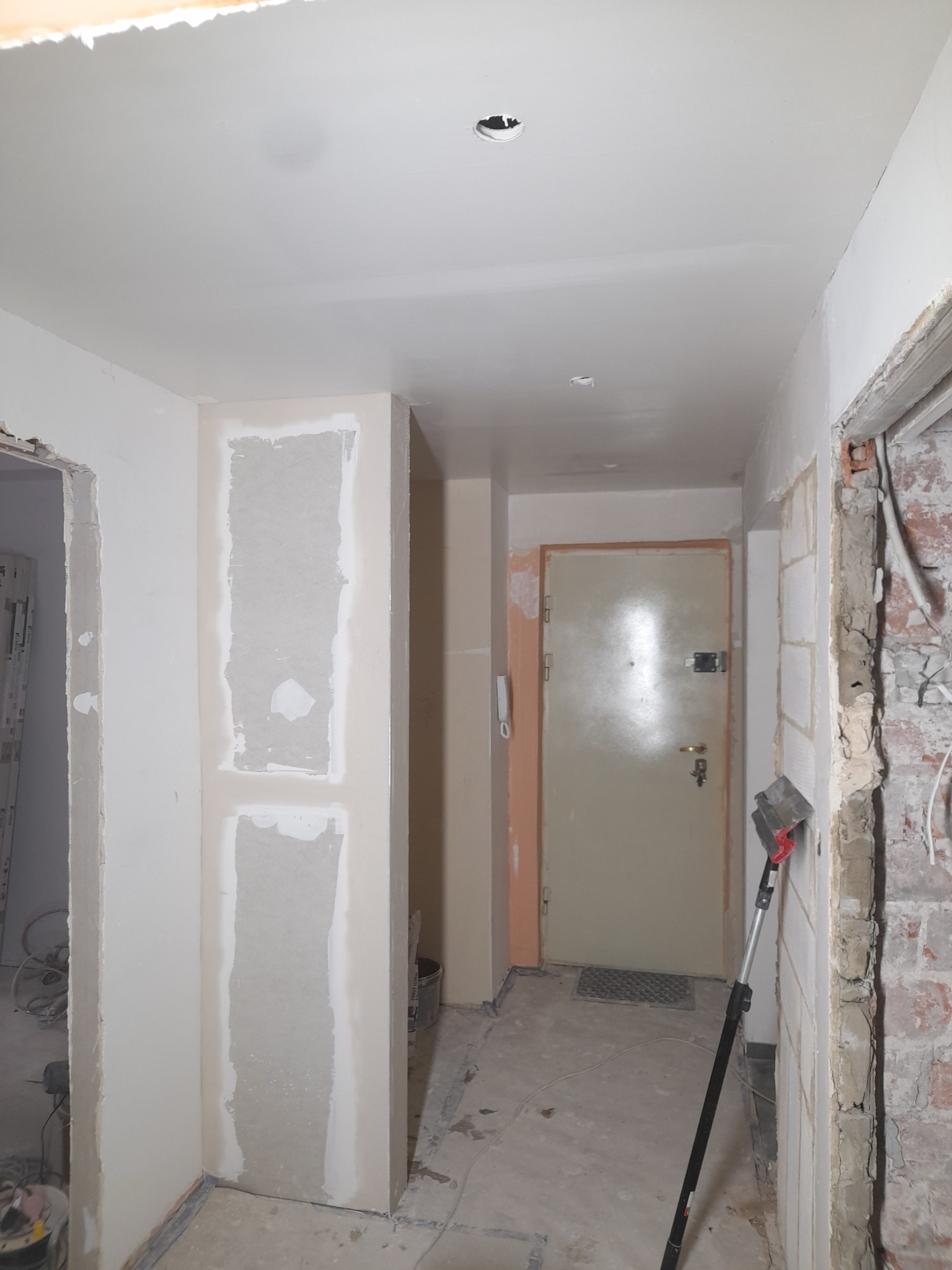 Korytarz w trakcie prac. Widoczne ślady po szpachlowaniu ścian, sufitu i obramowań drzwi. Na podłodze widoczne ślady po remoncie. Oprawa oświetleniowa bez żarówki.