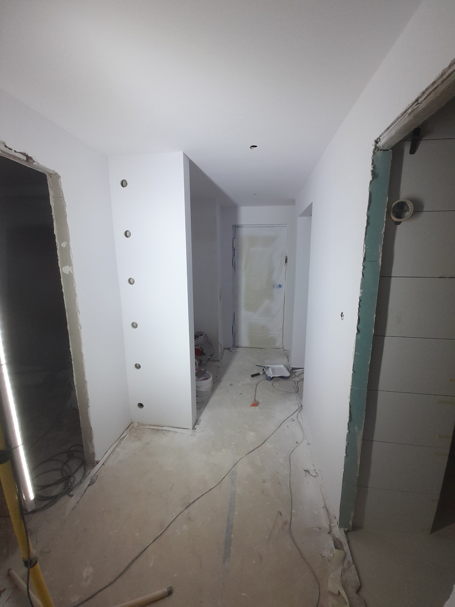 Korytarz w trakcie prac wykończeniowych: ściany pomalowane na biało, widoczne otwory na oświetlenie, podłoga przygotowana do dalszych prac. Surowy stan.