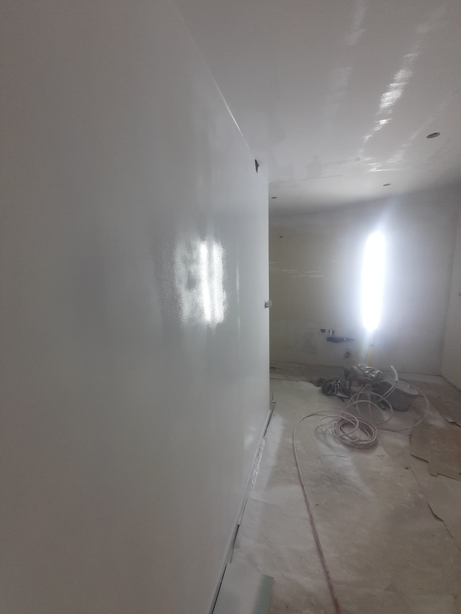 Świeżo pomalowane, białe, błyszczące ściany w pomieszczeniu. Widoczne narzędzia malarskie i niedoskonałości podłogi, sugerujące trwający remont. Oświetlenie halogenowe podkreśla gładkość powierzchni.