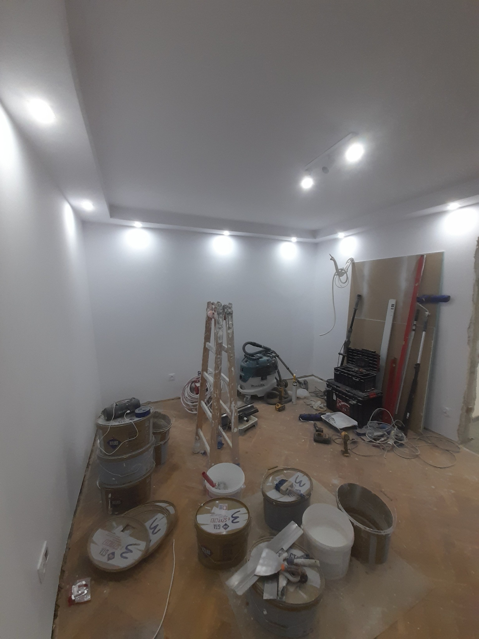 Pokój w trakcie prac wykończeniowych: białe ściany, sufit z oświetleniem, drabina, wiadra z farbą, narzędzia malarskie i płyta gipsowa. Widok z góry na rozgardiasz.