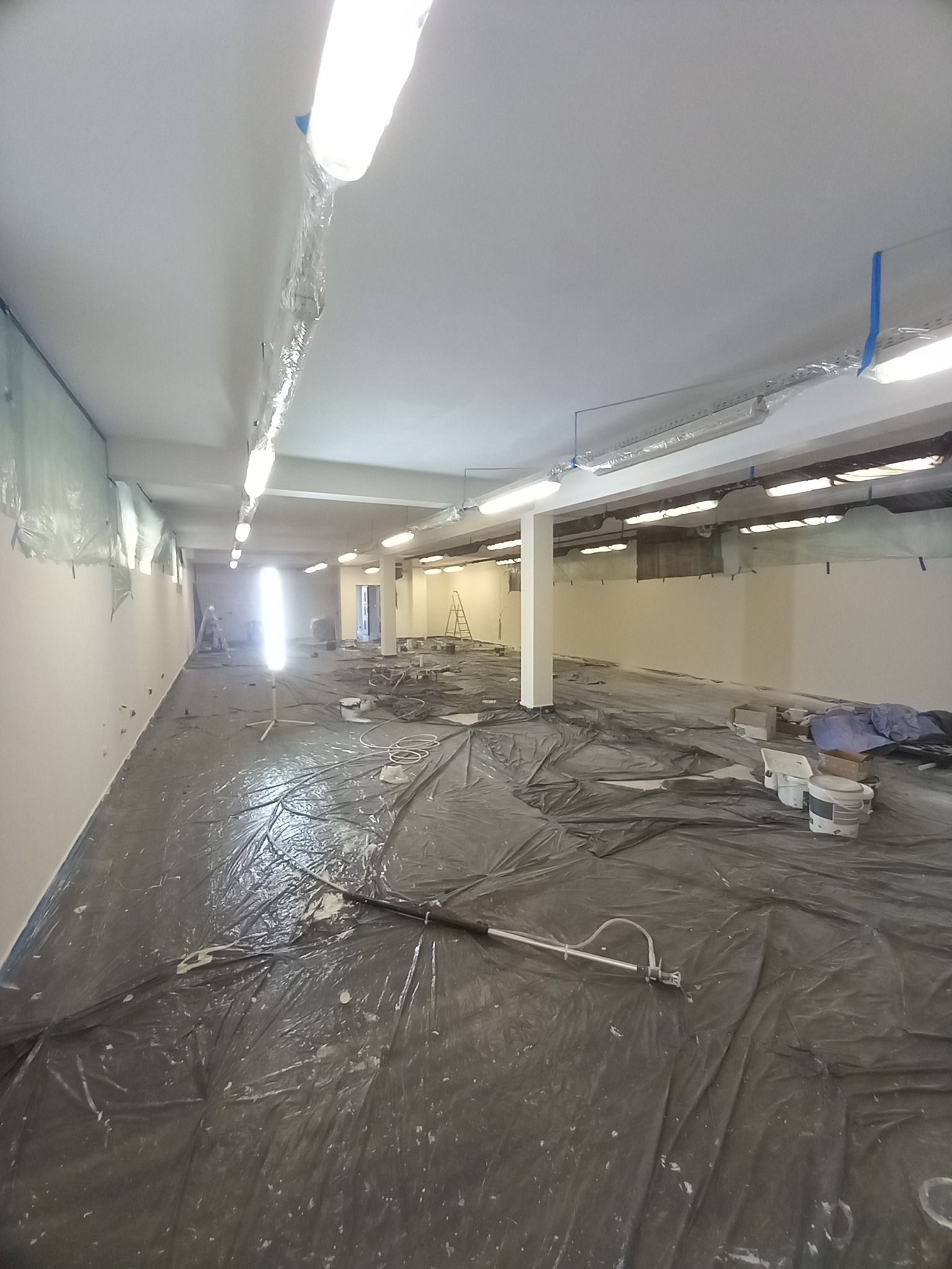 Wnętrze w trakcie malowania: podłoga zabezpieczona folią, ściany świeżo pomalowane na biało, widoczny sprzęt malarski i drabina w tle. Oświetlenie fluorescencyjne.