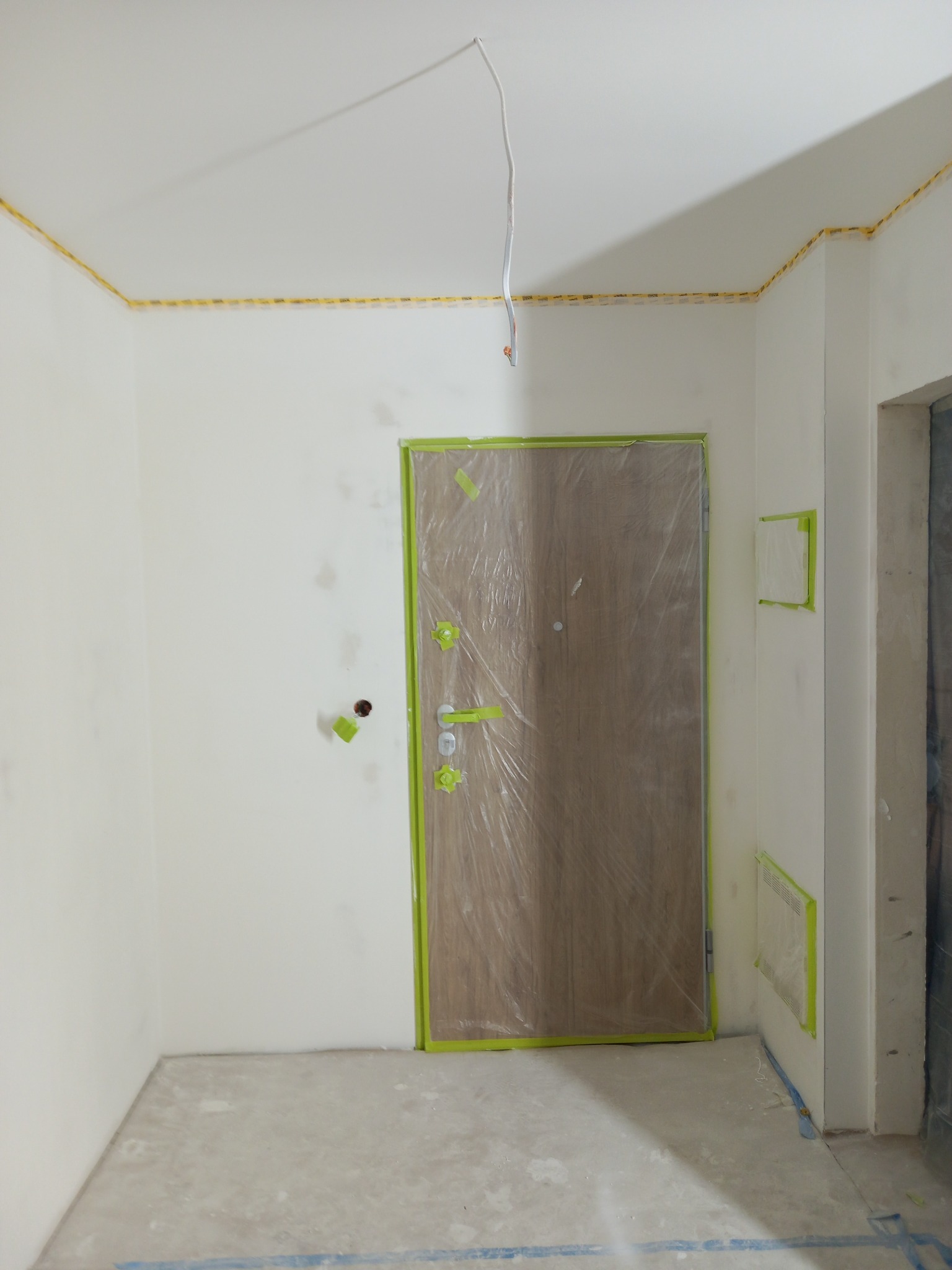Przygotowania do malowania: drzwi zabezpieczone folią i taśmą, ściany świeżo pomalowane na biało, widoczne wystające kable.