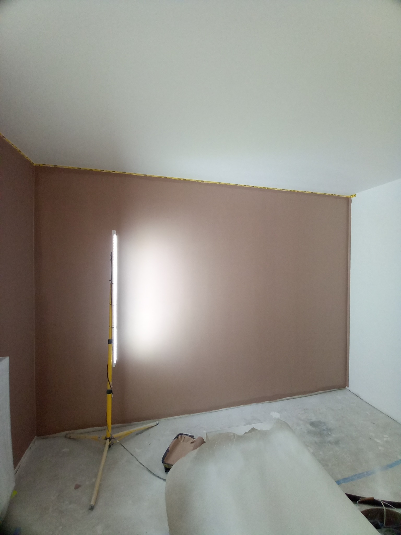 Pokój po szpachlowaniu ścian na brązowo, z taśmą malarską przy suficie i lampą budowlaną na statywie. Widoczny fragment podłogi i resztki materiałów.