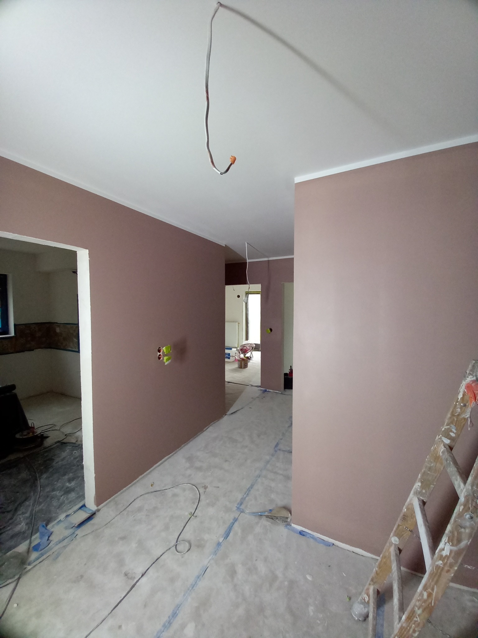 Wnętrze w trakcie malowania: ściany w odcieniach różu, zabezpieczona podłoga, drabina malarska z boku, wystające kable. Widok na dalsze pomieszczenia.