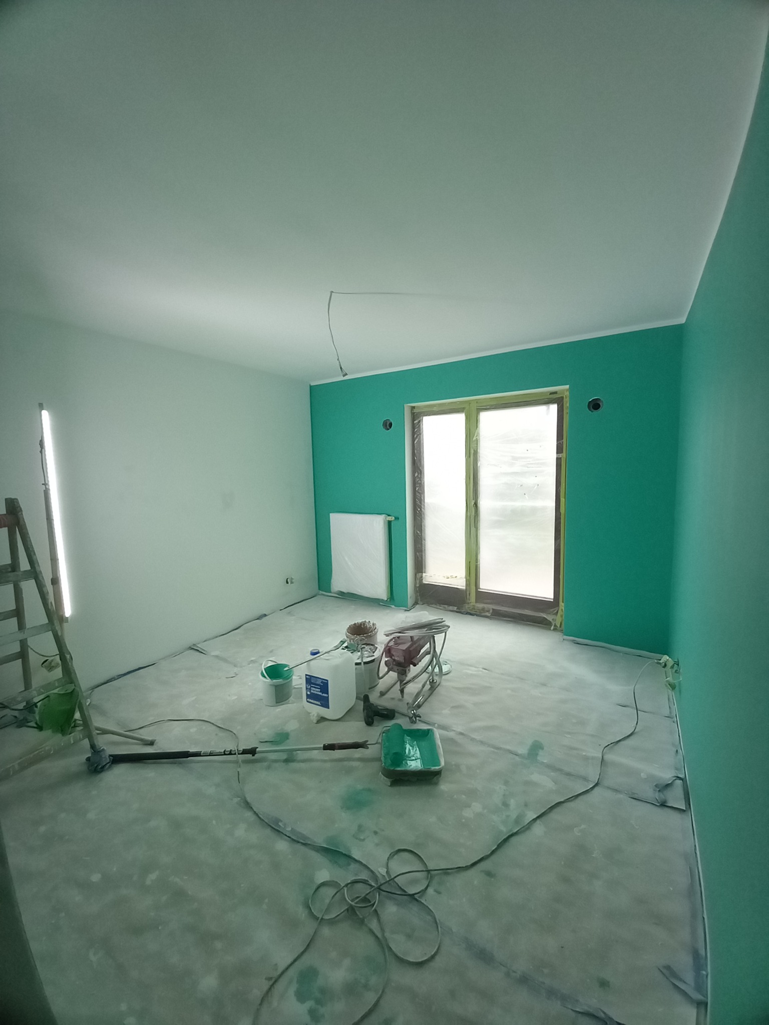 Pokój po malowaniu natryskowym na dwa kolory: biały sufit i turkusowe ściany. Widoczne narzędzia malarskie i zabezpieczona folią podłoga.