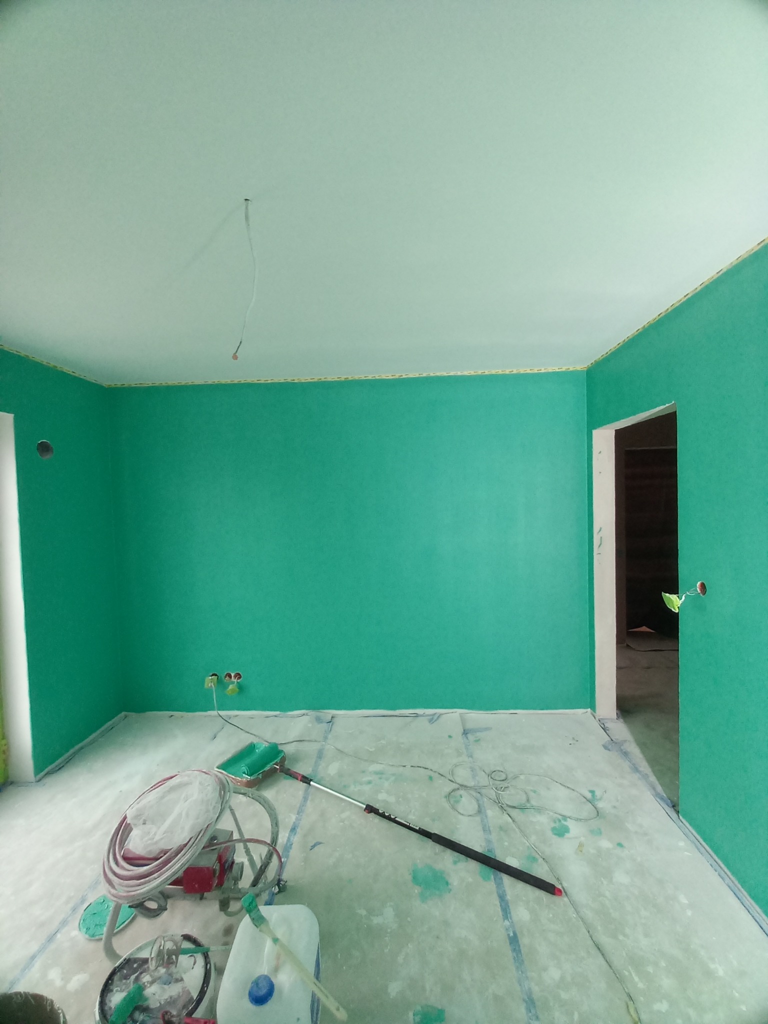 Świeżo pomalowane na zielono wnętrze pokoju z zabezpieczoną podłogą i sprzętem malarskim. Widoczne gniazdka elektryczne i otwór w ścianie.