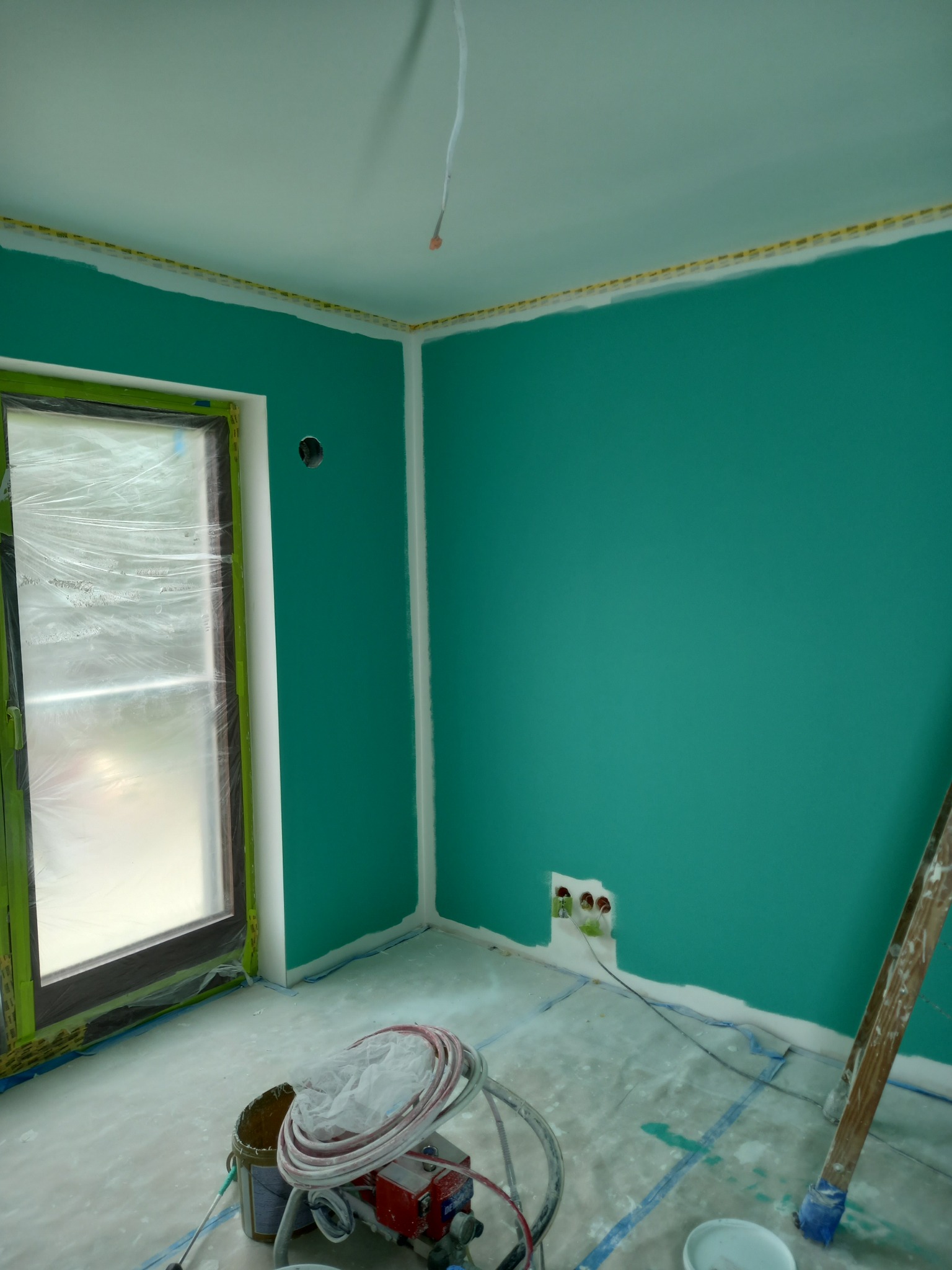Świeżo pomalowane na turkusowo wnętrze z zabezpieczonym oknem, widoczne narzędzia malarskie i drabina. Przygotowane do dalszych prac wykończeniowych.