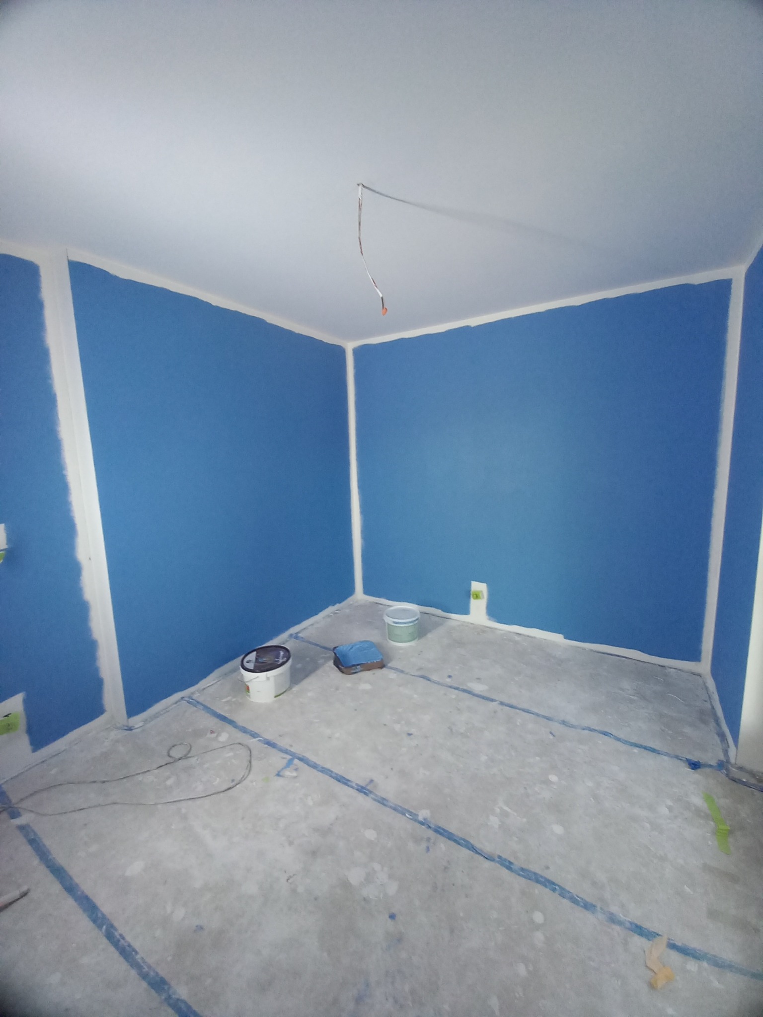 Pokój po malowaniu na niebiesko, z białymi listwami i zabezpieczoną podłogą. Widoczne puszki z farbą i kabel elektryczny wystający ze ściany.