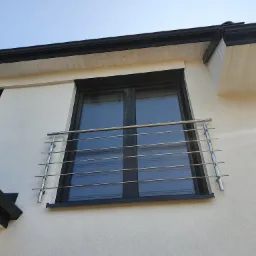 Zewnętrzna, pozioma, metalowa barierka ochronna, zamontowana przed oknem na elewacji budynku z czarną ramą okienną i czarnym orynnowaniem.