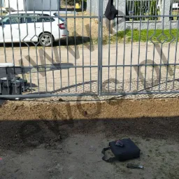 Szara, metalowa brama wjazdowa z automatyką, widoczna na tle podwórka z samochodami; ziemia wokół bramy świeżo przekopana, obok leżą narzędzia w czarnej torbie.