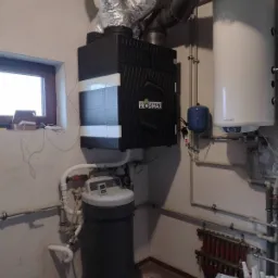 Czarna jednostka rekuperacyjna PRODMAX z srebrnymi przewodami wentylacyjnymi, obok biały zasobnik ciepłej wody, niebieskie naczynie przeponowe, grzejnik podłogowy i zmiękczacz wody w kotłowni.