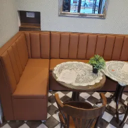 Wnętrze kawiarni z narożną, brązową kanapą ze skóry ekologicznej, okrągłymi stolikami z koronkowymi serwetami i drewnianymi krzesłami na czarno-białej podłodze.