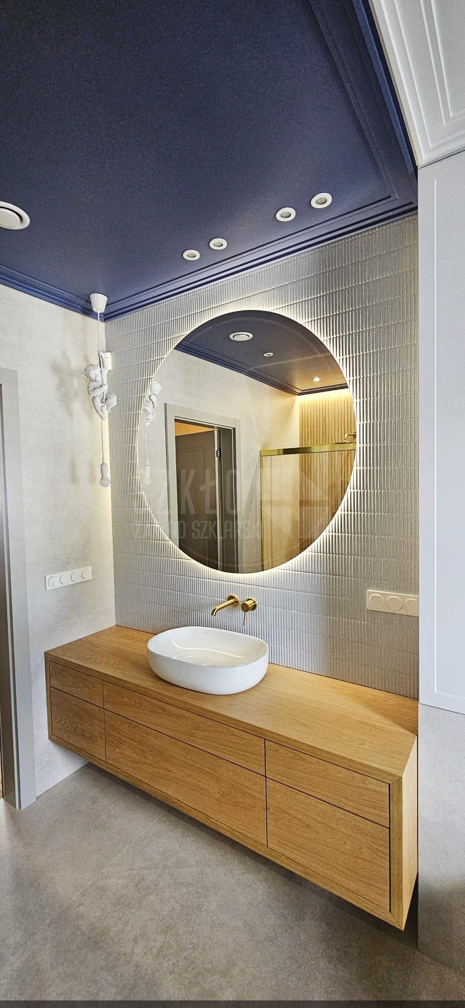 Nowoczesna łazienka z okrągłym lustrem LED, drewnianą szafką pod umywalką i designerską lampą sufitową. W lustrze odbija się kabina prysznicowa.