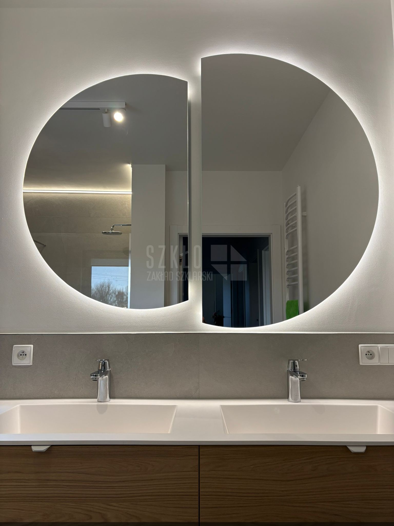 Okrągłe lustro z podświetleniem LED nad umywalką w łazience w Szczecinie. Nowoczesny design, szara ściana, drewniane szafki pod umywalką. Widoczne detale wykończenia.