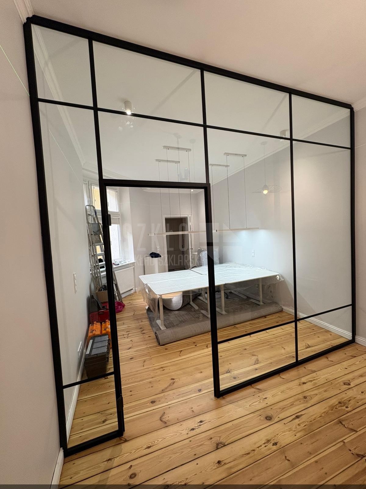 Nowoczesna, przeszklona ścianka działowa z czarnymi profilami, oddzielająca przestrzeń biurową z drewnianą podłogą. Widoczne biurka i drabina w tle.