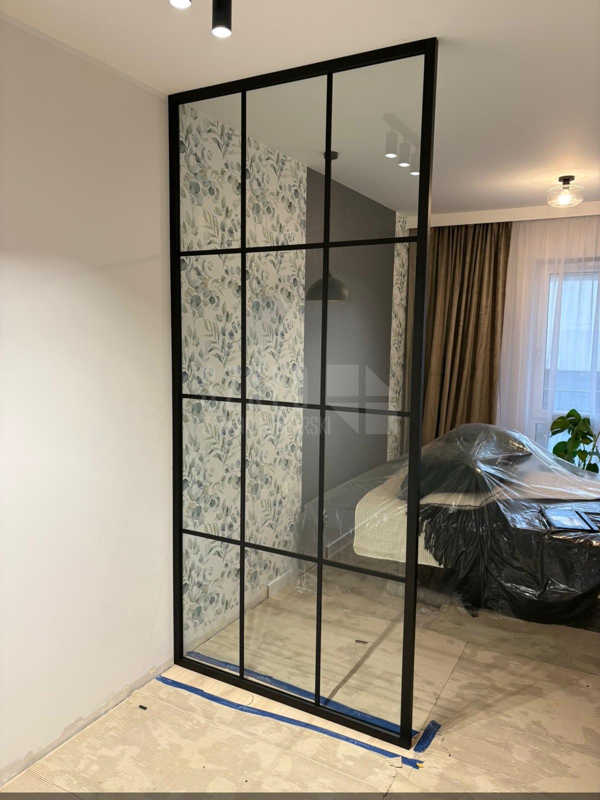Szklana ścianka działowa z czarnymi profilami, oddzielająca pokój z tapetą w liście od sypialni z łóżkiem przykrytym folią ochronną. Widoczne oświetlenie sufitowe.