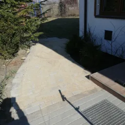 Świeżo ułożona ścieżka z kostki brukowej, zasypana piaskiem, prowadząca do domu z widoczną studzienką odpływową i zielonym iglakiem obok.