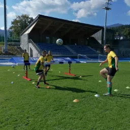 Trening piłkarski na boisku w Karpaczu: grupa młodzieży w żółto-zielonych koszulkach ćwiczy podbijanie piłki głową nad niską przeszkodą, w tle trybuny i góry.