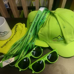 Superwypoczynek.pl - Zestaw promocyjny: zielone czapki, okulary, smycze z napisem 'Obozy sportowe, kolonie' oraz kubek i woreczek z logo 'superwypoczynek.pl'.