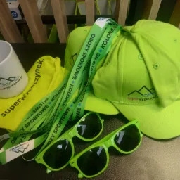 Zestaw promocyjny: zielone czapki, okulary, smycze z napisem 'Obozy sportowe, kolonie' oraz kubek i woreczek z logo 'superwypoczynek.pl'.