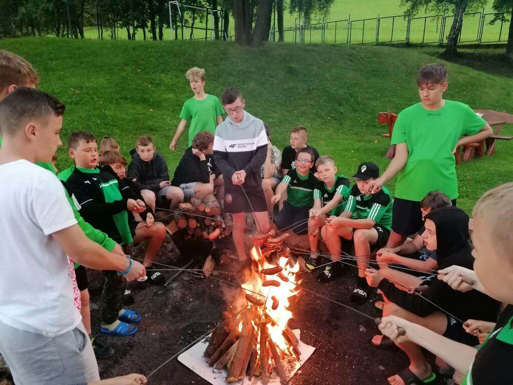 Grupa chłopców w Sosnowcu siedzi wokół ogniska, piekąc kiełbaski na patykach podczas wieczornego spotkania na zielonym terenie.