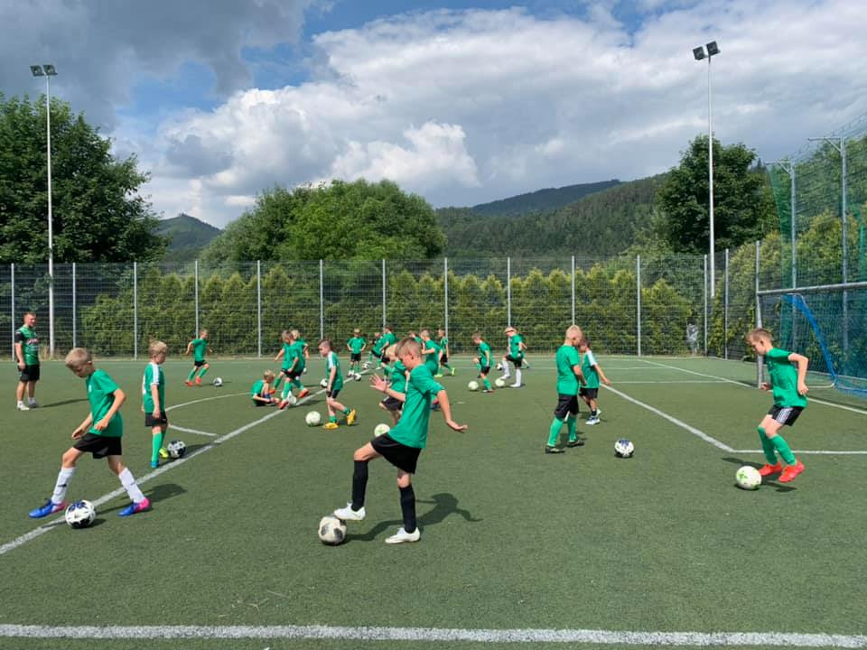 Grupa dzieci w zielonych koszulkach trenuje piłkę nożną na zielonym boisku z białymi liniami, w tle drzewa i góry pod błękitnym niebem z chmurami.