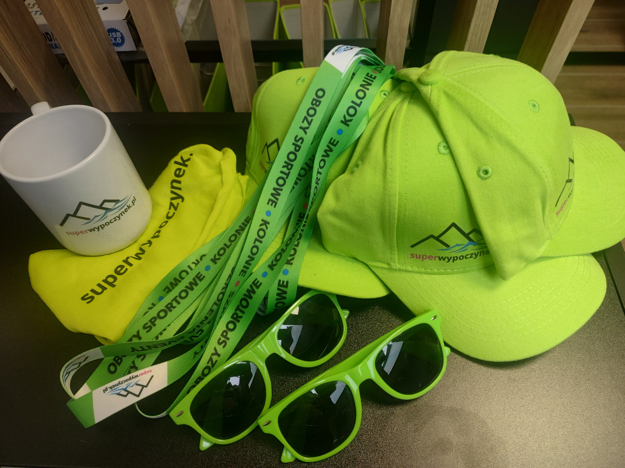 Zestaw promocyjny: zielone czapki, okulary, smycze z napisem 'Obozy sportowe, kolonie' oraz kubek i woreczek z logo 'superwypoczynek.pl'.