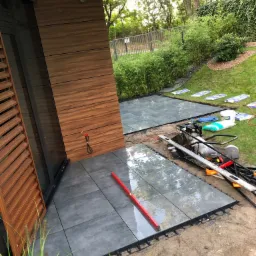 Montaż szarych płyt tarasowych na regulowanych wspornikach, widoczny poziomica i narzędzia, trawa i roślinność w tle.