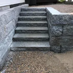 Schody zewnętrzne z szarego kamienia prowadzące do domu, otoczone kamiennym murem oporowym i żwirem.