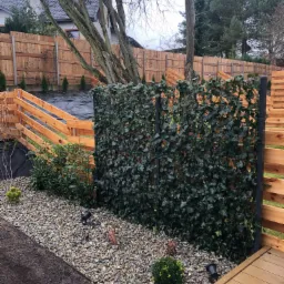 Nowoczesny ogród z drewnianym ogrodzeniem, panelami z bluszczu na metalowych słupkach, kamienną rabatą i fragmentem tarasu kompozytowego w pochmurny dzień.