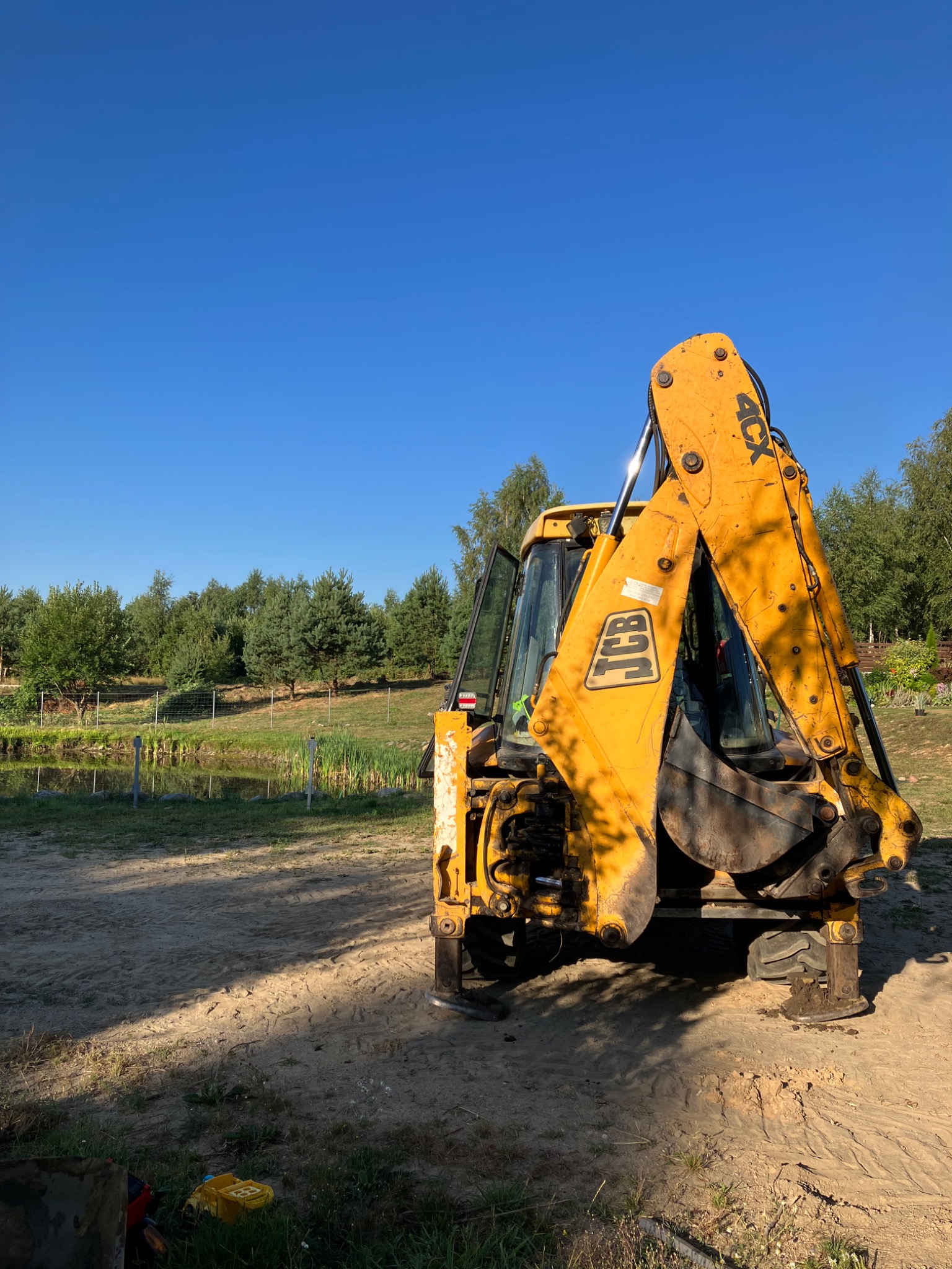 Żółta koparko-ładowarka JCB 4CX na piaszczystym terenie, w tle małe jezioro i drzewa pod błękitnym niebem.