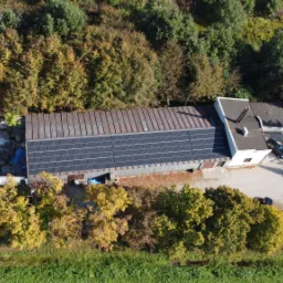 Widok z drona na budynek z panelami fotowoltaicznymi na dachu, otoczony jesiennymi drzewami i zabudowaniami.